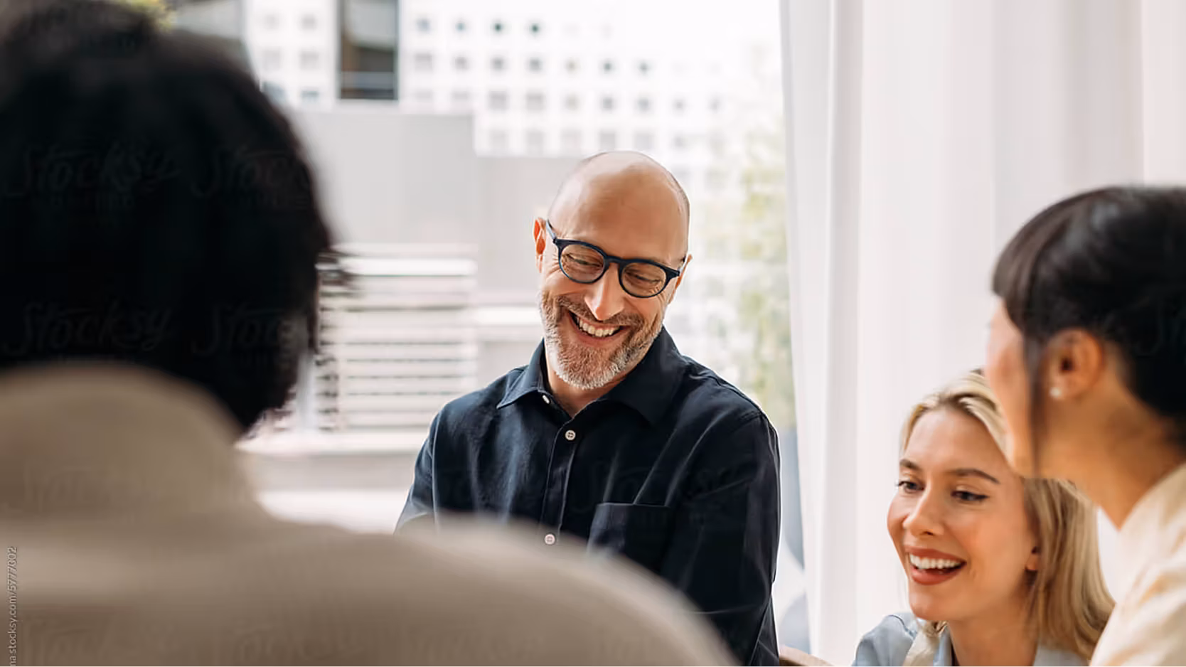 A group of professionals smiling and laughing during a casual business meeting near a window, with a bald man in glasses at the center of the image.